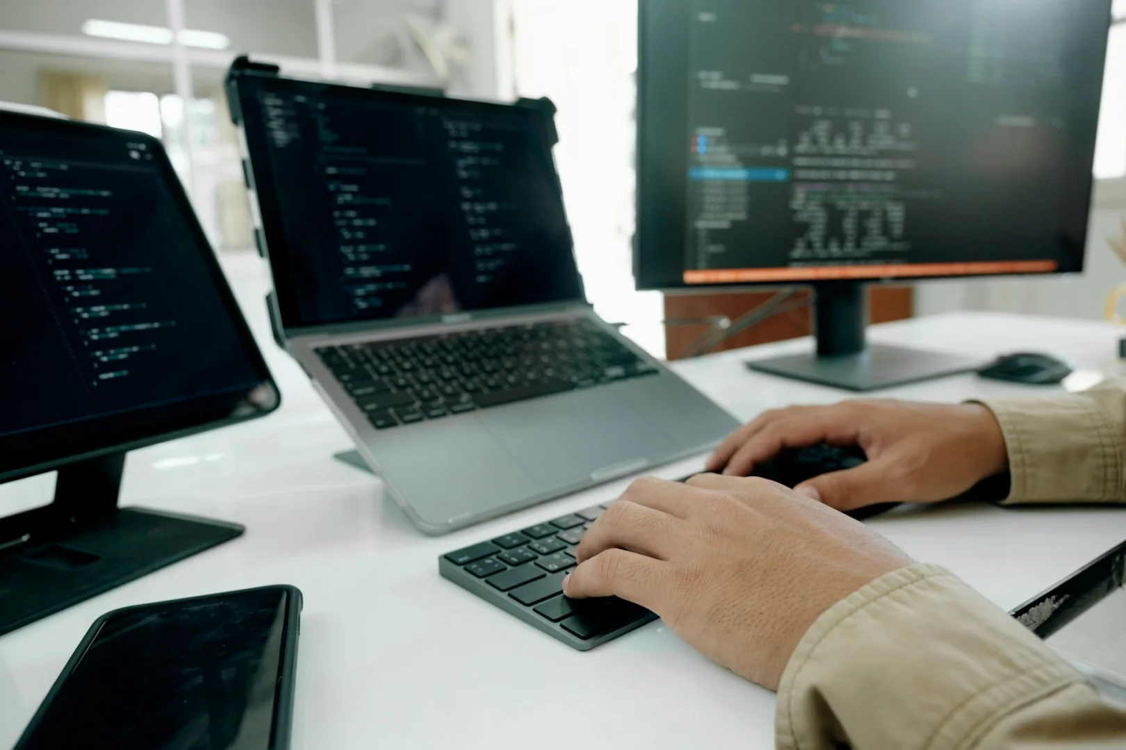 Das Foto der Hände eines Softwaredevelopers auf der Tastatur vor 3 Geräten unterstreicht unterschiedliche Ansätze von Websites